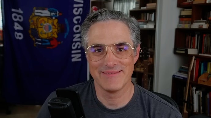 Retrato em close do diretor de jogos Josh Sawyer sorrindo de forma tímida durante transmissão em seu canal de vídeo.